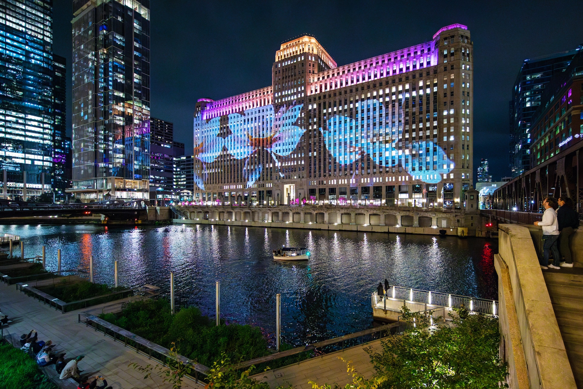 A nighttime view of a large building with colorful lighting on its facade, overlooking a river with a boat and cityscape in the background.