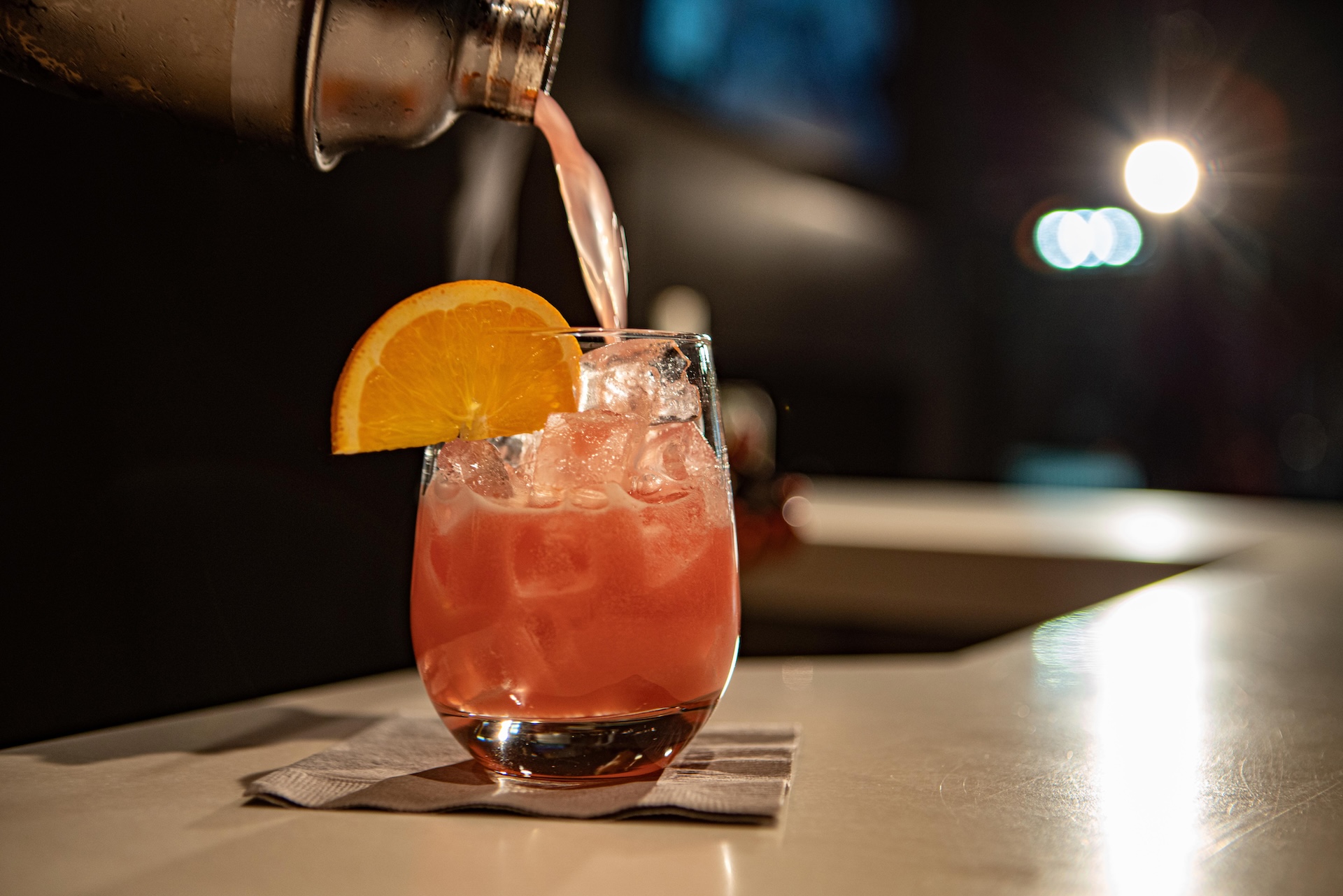 A pink cocktail being poured into a glass with ice and an orange slice.